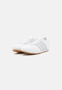 White athletic shoes made of synthetic leather, featuring silver stripes, textured detailing on the toe, and a gum rubber outsole.