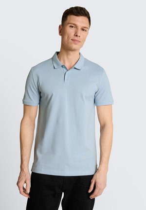 Mężczyzna ubrany w jasnoniebieską koszulkę polo z krótkim rękawem i czarne spodnie, stojący na jednolitym, jasnoszarym tle.