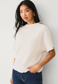 Helle, beigefarbene, strukturierte Bluse mit hohem Kragen und kurzen Ärmeln, die mit gewellten Kanten versehen ist, kombiniert mit dunkelblauen Jeans.