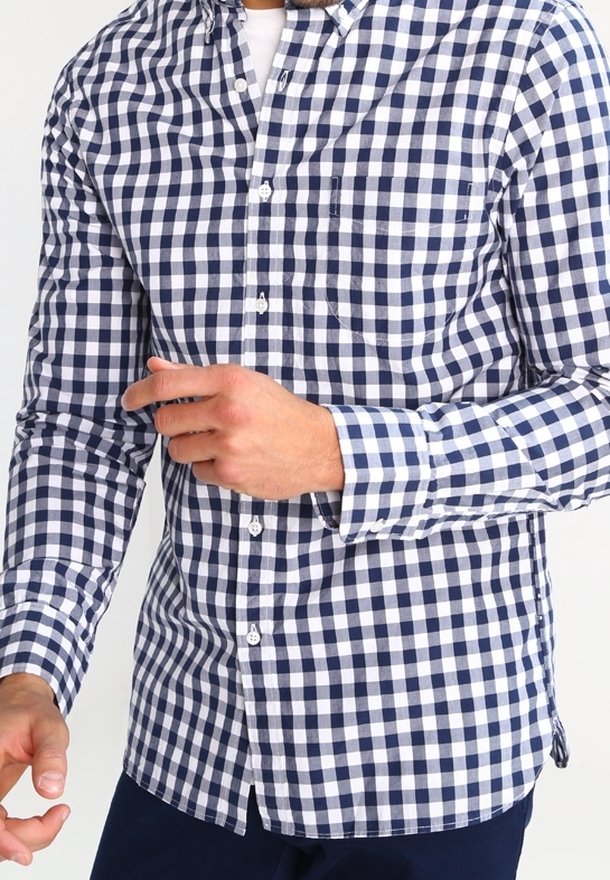 Homme portant une chemise à carreaux bleue et blanche boutonnée, la main gauche légèrement levée sur un fond uni.