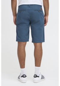 Bermudas de algodón azul con corte recto, dos bolsillos traseros y cierres de botón. Llevadas con zapatillas blancas y calcetines visibles hasta el tobillo.