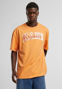 Orange bomulds T-shirt med "KANI" logo i hvid og rød, afslappet pasform, korte ærmer og rund halsudskæring. Bæret med denimshorts.