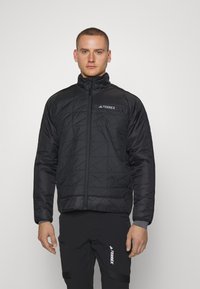 Chaqueta negra acolchada con un cuello alto, cremallera frontal y logotipo en la parte superior izquierda. Lleva puesta sobre pantalones negros a juego. Tela suave y duradera.