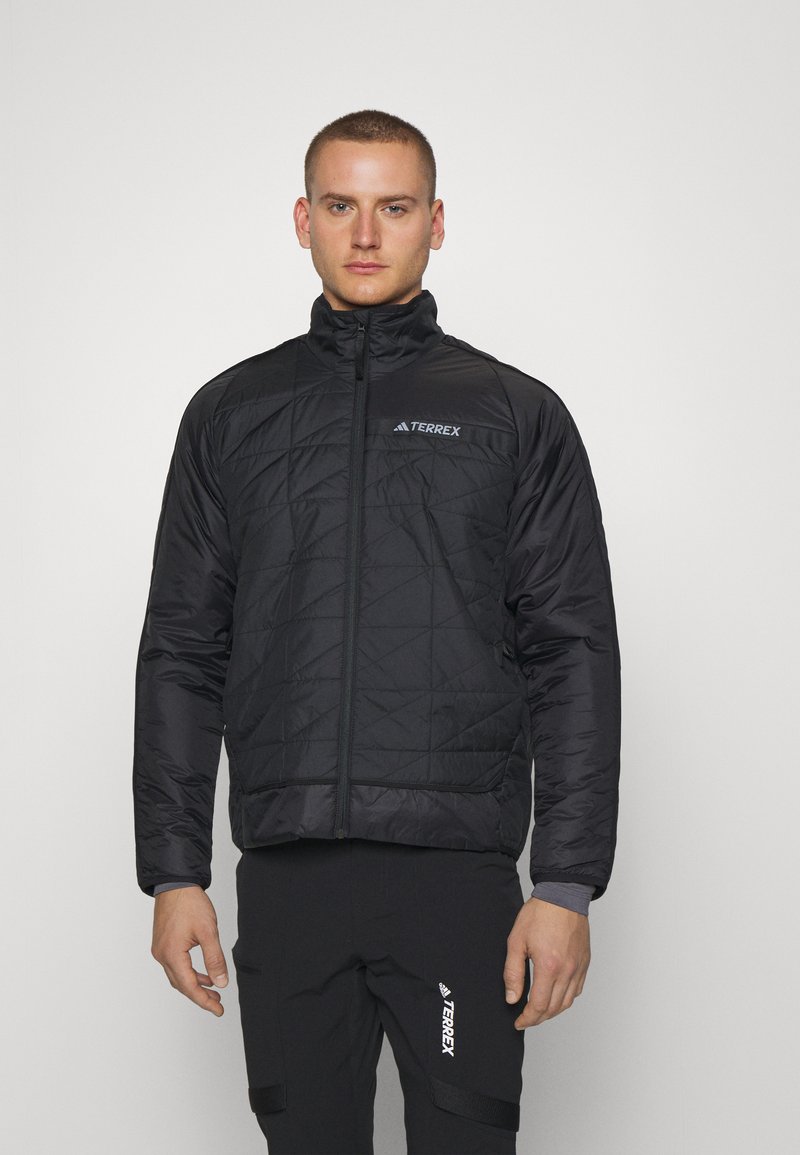 Chaqueta negra acolchada con un cuello alto, cremallera frontal y logotipo en la parte superior izquierda. Lleva puesta sobre pantalones negros a juego. Tela suave y duradera.
