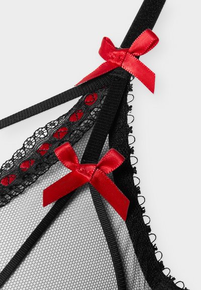 Λεπτομέρεια από μαύρη δαντελένια εσώρουχα με διάφανο ύφασμα από δίχτυ και κόκκινα σατέν φιόγκους σε λεπτές μαύρες τιράντες πάνω σε λευκό φόντο.