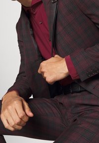 Traje de tartán burdeos con ribete negro, que presenta un corte ajustado, solapas con muesca y una camisa a juego en color burdeos. Cinturón de cuero negro visible.