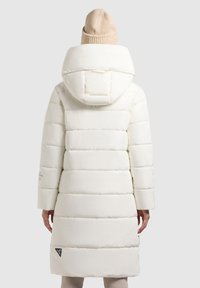 White padded coat with a large hood, featuring horizontal stitching, long sleeves, and a side logo detail. Accompanied by a beige knitted beanie.