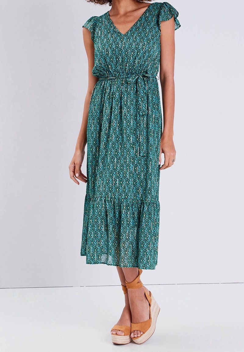 Femme portant une robe midi bleu sarcelle à motifs, avec des manches courtes et une ceinture à nouer à la taille, assortie à des sandales compensées camel.