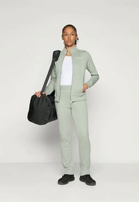 Hele roheline spordikostüüm, millel on lukuga jakki ja sobivad püksid. Isik hoiab käes musta duffel-kotti. Mustad jalanõud completeerivad riietust.