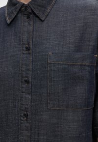 Gros plan du devant d’une chemise en denim foncé avec des boutons noirs, un col pointu, et une poche poitrine rectangulaire avec une couture orange.