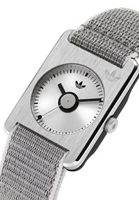 Kvadratinis sidabrinis riešo laikrodis su pilku pintu dirželiu, juodais valandų ir minučių rodyklėmis bei Adidas trèfoil logotipu ant ciferblato ir korpuso.