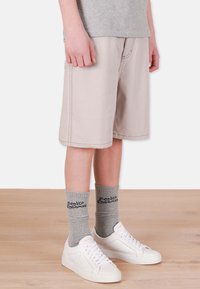 Beige denimshorts med synliga sömmar, i kombination med grå strumpor med text och vita sneakers med perforerat mönster och snörning.