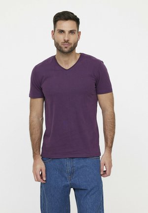 T-shirt violet à col en V en tissu doux avec des manches courtes, présenté avec un jean en denim bleu. Détail de logo minimal sur la poitrine.