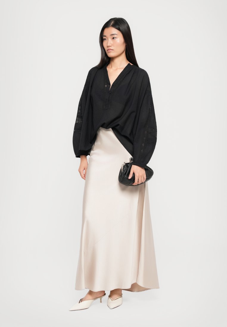 Blouse noire ample à manches longues, assortie à une jupe maxi en satin crème. Les accessoires comprennent un petit sac à main noir et des ballerines à bout pointu.