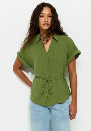 Woman with long curly hair wearing a short-sleeve green button-up shirt with a waist tie and light blue jeans, standing against a plain background.