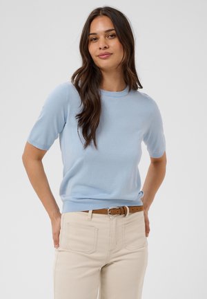 Femme aux longs cheveux foncés portant un pull à manches courtes bleu clair, un pantalon beige et une ceinture marron, debout, les mains dans le dos.