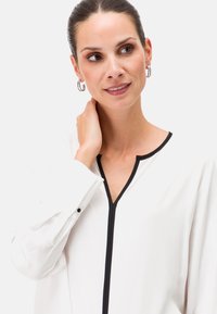 Weiße Bluse mit V-Ausschnitt, schwarzem Saum und langen Ärmeln, mit geknöpften Manschetten und einer glatten Stoffoberfläche.