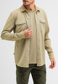 Khakifarbene Knopfleiste mit zwei Brusttaschen, langen Ärmeln und geradem Saum, darüber getragen ein hellbeiges Rundhals-T-Shirt.