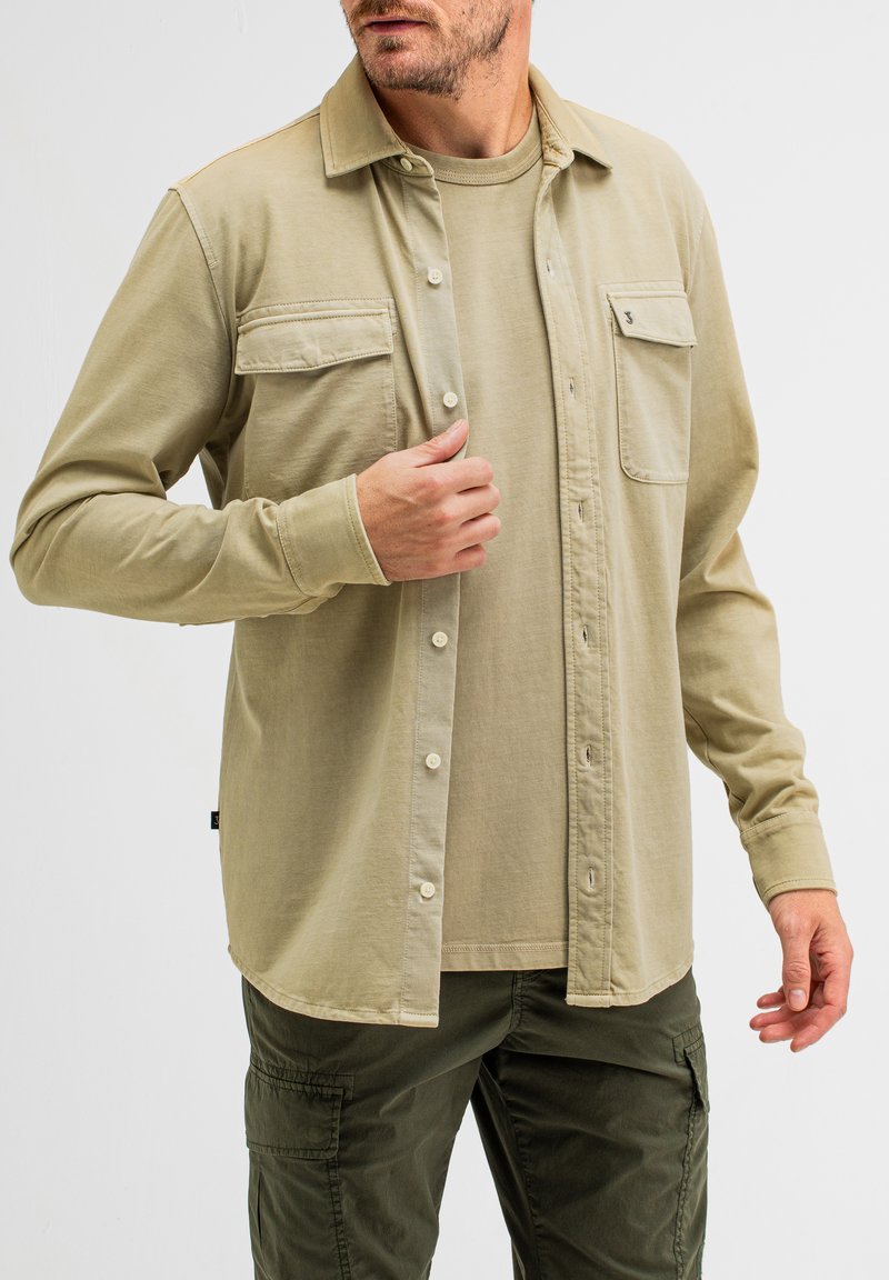 Khakifarbene Knopfleiste mit zwei Brusttaschen, langen Ärmeln und geradem Saum, darüber getragen ein hellbeiges Rundhals-T-Shirt.