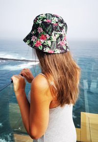 Cappello a secchiello nero con motivi floreali rosa e verdi tropicali, con logo. Modella in piedi accanto a una ringhiera di vetro con vista sull'oceano.