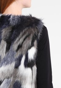 Faux fur-väst med en blandning av grå, vit och svarta nyanser, med en mjuk textur och ett naturligt mönster, bärs över en svart topp.