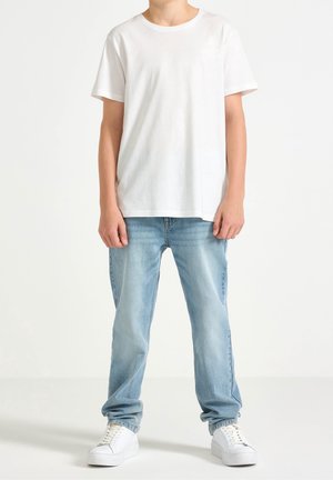 Biały bawełniany t-shirt z krótkimi rękawami, w zestawieniu z jasnoniebieskimi dżinsami. Model ma na sobie białe trampki. Prosty, casualowy design.