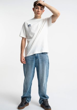 Młody mężczyzna w białej koszulce z nadrukiem, luźnych niebieskich dżinsach i czarnych tenisówkach, stoi z jedną ręką na głowie na jednolitym tle.