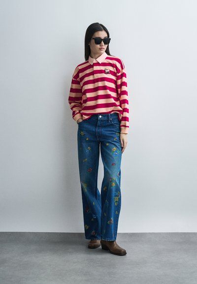 Γυναίκα που φοράει μαύρα γυαλιά ηλίου, μακρυμάνικο πουκάμισο με ρίγες σε κόκκινο και ροζ χρώμα με κεντημένο ελέφαντα, τζιν παντελόνι με κεντήματα λουλουδιών σε μπλε απόχρωση και καφέ μπότες.