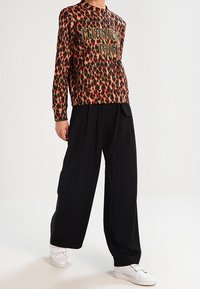 Personne portant une chemise à manches longues imprimée léopard avec le texte "Midnight Run", un pantalon large noir et des baskets blanches, debout sur un sol blanc.