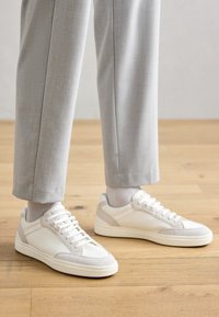 Weiße Sneaker aus glattem Synthetikmaterial mit hellbeigen Akzenten. Mit runder Zehenpartie, flacher Sohle und Schnürdesign. Getragen mit hellgrauen Hosen.