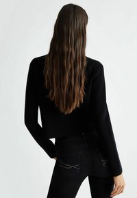 Kvinde med langt brunt hår iført en sort ribstrikket sweater og sorte jeans med rhinsten-detaljer på baglommerne, stående mod en hvid baggrund.