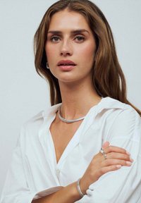 Silverhalsband med en slät, rund design. Modellen bär en vit skjorta med knappar. Ett enkelt silverarmband och en ring syns på armen.