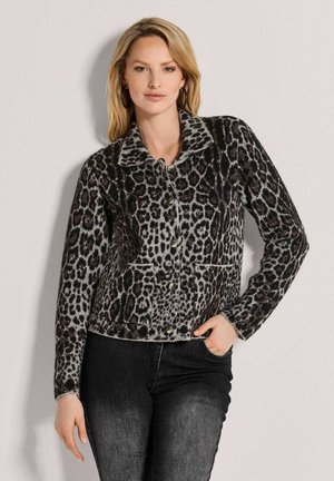 Frau mit blonden Haaren, die eine geknöpfte, langärmelige Jacke mit Leopardenmuster und dunkle Jeans trägt, steht vor einer einfachen, hellen Wand.