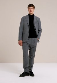 Szary garnitur w prążki z dopasowaną marynarką i pasującymi spodniami, w połączeniu z czarnym topem o wysokim kołnierzu. Teksturowany materiał i elegancki design.