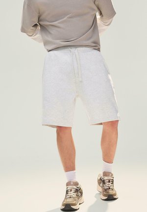 Persona che indossa pantaloncini grigio chiaro con coulisse, scarpe da ginnastica beige e bianche, calzini bianchi e una maglietta taupe a maniche corte, in piedi con le gambe divaricate.