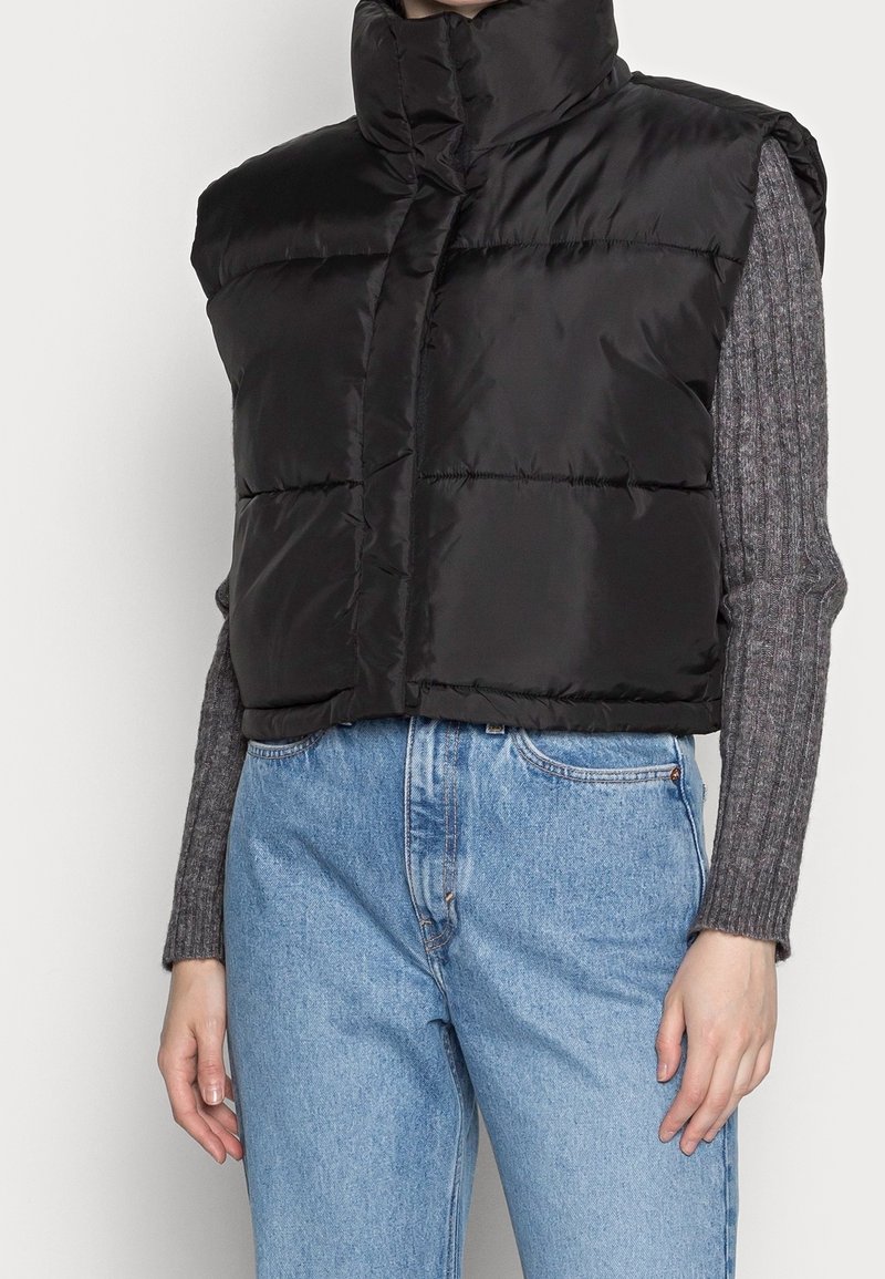 Black puffer vest with high collar, sleeveless design, and shiny texture, worn over a gray ribbed sweater and light blue jeans.