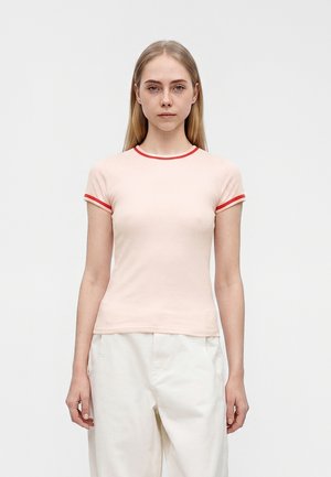 Ung kvinde med langt blondt hår iført en lyserød tætsiddende t-shirt med røde kanter og løse hvide bukser, stående mod en ensfarvet hvid baggrund.