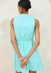 Un vestito senza maniche, lungo fino al ginocchio, di colore azzurro chiaro con un motivo ad occhiello, cintato in vita e con una chiusura posteriore a bottoni.