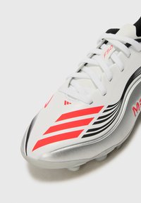Witte voetbalschoenen met rode en zwarte strepen, gemaakt van nylon materiaal, met zilveren accenten en witte veters. Heeft een slank, gestroomlijnd ontwerp.