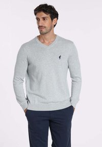 Pull en maille gris avec un col en V, des poignets et un ourlet côtelés, arborant un petit emblème de cheval bleu marine sur la poitrine gauche. Texture lisse, coupe décontractée.