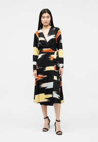 Γυναίκα που φοράει μαύρο φόρεμα τύπου wrap με μακριά μανίκια και αφηρημένα σχέδια με πινελιές πορτοκαλί, κίτρινου και λευκού, συνδυασμένο με μαύρα τακούνια.