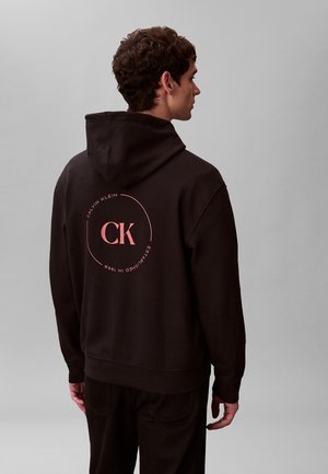 Hombre de espaldas con una sudadera negra con capucha que tiene un logo circular rosa de Calvin Klein en la parte posterior, sobre un fondo gris liso.