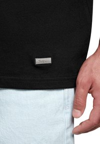 Black t-shirt with a textured fabric, featuring a small metallic logo tag at the hem, paired with light blue denim shorts.