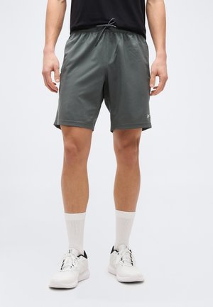 Hombre con pantalones cortos deportivos grises con cordón, calcetines blancos hasta la pantorrilla y zapatillas blancas, de pie contra un fondo liso.