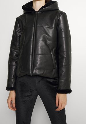 Person trägt eine schwarze gesteppte Jacke mit Kapuze und Reißverschluss vorne, kombiniert mit schwarzen, strukturierten Hosen, vor einem einfarbigen hellen Hintergrund.