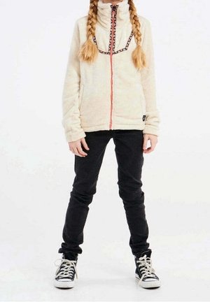 Giacca in pile color crema con colletto alto e cerniera, caratterizzata da un motivo geometrico colorato sul petto. Abbinata a pantaloni slim fit neri e sneakers.