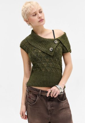 Junge Person mit kurzen blonden Haaren, die ein grünes gestricktes schulterfreies Oberteil und braune Hose trägt, posiert mit einer Hand in der Nähe der Taille.