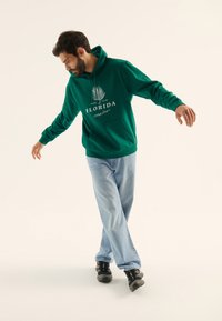 Sudadera verde con capucha, bolsillo frontal y texto gráfico en blanco, combinada con jeans azul claro y zapatillas negras, de pie sobre un pie.