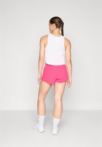 Weißes geripptes Tanktop und leuchtend pinke Sportshorts mit figurnaher Passform. Modell von hinten zu sehen, trägt weiße Socken und Schuhe.