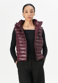 Bordeaux puffer vest med høy krage, quiltet design og glidelås. Brukt over en svart topp, som viser en figurvennlig silhuett.